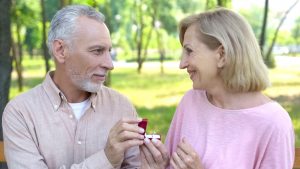A senior couple sits in a park. He offers her a diamond ring.